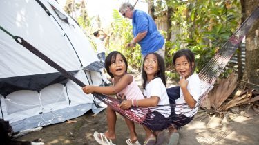 20 Jahre ShelterBox. Drei Kinder auf den Philippinen. Im Hintergrund sieht man ein ShelterBox Zelt, das ihre Familien nach dem Typhoon als Katastrophenhilfe erhalten haben